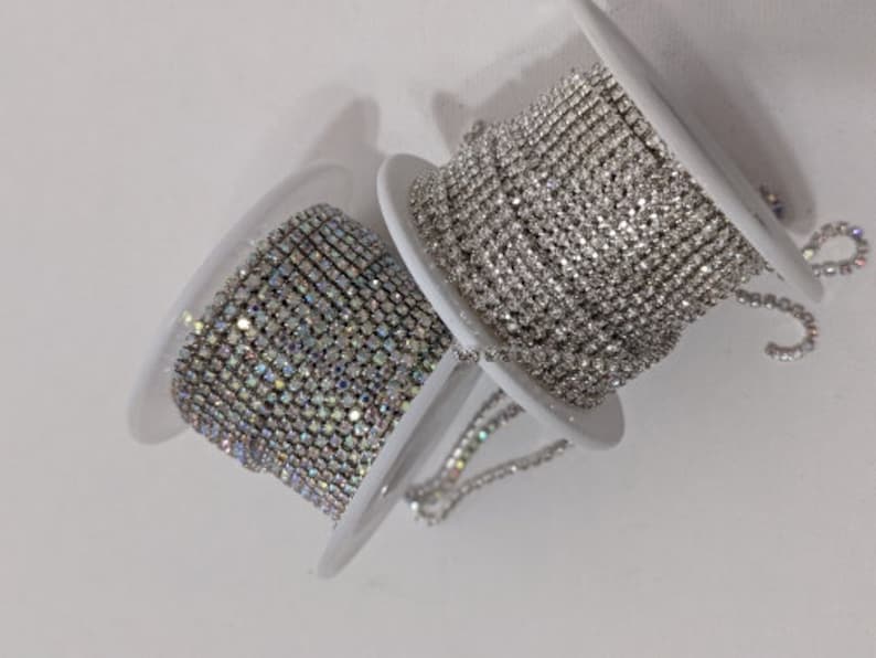 May include: Two spools of rhinestone chain. One spool features clear rhinestones, while the other has iridescent rhinestones. The spools are white and are set against a white background. The rhinestone chain is likely used for jewelry making or embellishments.