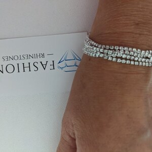 May include: A sparkling rhinestone bracelet with multiple strands of clear, round rhinestones. The bracelet is worn on a wrist, and a white card with the word "FASHION" and the word "RHINESTONES" is visible.
