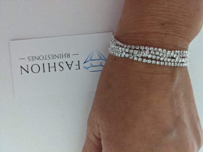 May include: A sparkling rhinestone bracelet with multiple strands of clear, round rhinestones. The bracelet is worn on a wrist, and a white card with the word "FASHION" and the word "RHINESTONES" is visible.