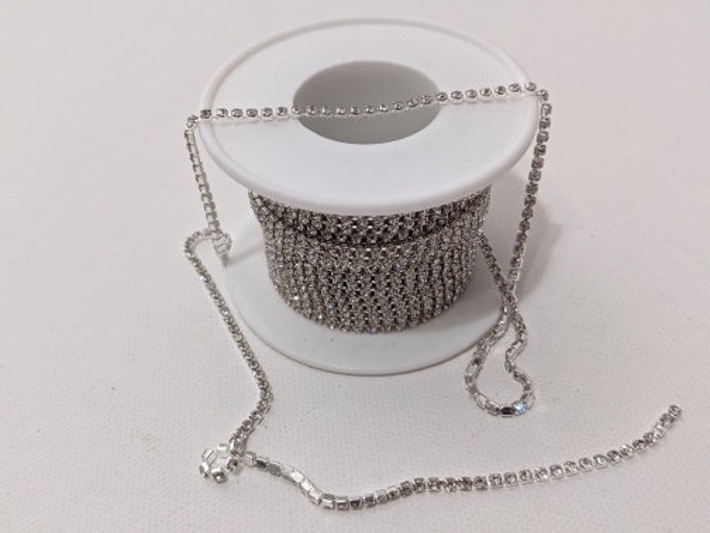 May include: A spool of silver rhinestone chain. The chain is coiled around a white plastic spool. A length of the chain extends from the spool, showcasing the sparkling, clear rhinestones. The background is a plain white surface.