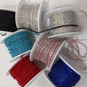 May include: Assortment of rhinestone chain rolls in various colors, including black, silver, iridescent, turquoise, pink, red, and blue. Each roll is neatly wound on a white spool, showcasing the sparkling texture of the rhinestones.