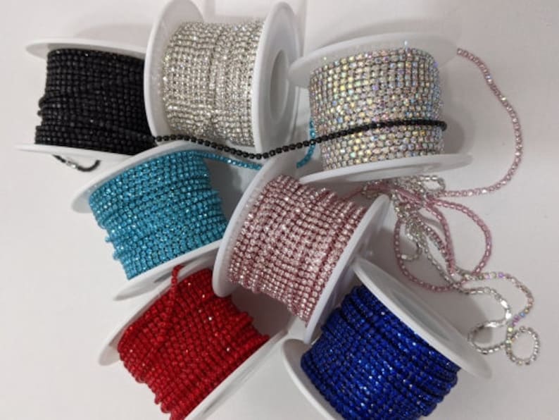 May include: Assortment of rhinestone chain rolls in various colors, including black, silver, iridescent, turquoise, pink, red, and blue. Each roll is neatly wound on a white spool, showcasing the sparkling texture of the rhinestones.