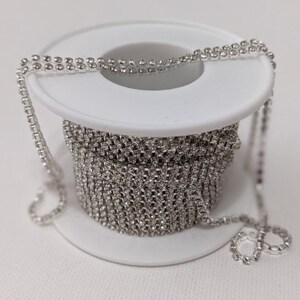 May include: A spool of silver rhinestone chain. The chain is wound around a white plastic spool. The chain is made of small, round rhinestones set closely together, creating a continuous, sparkling line. The chain is partially unwound.