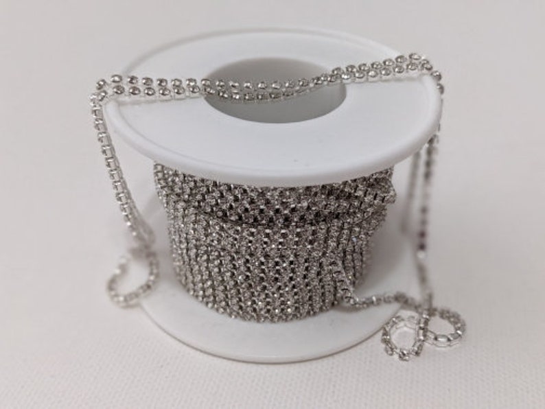 May include: A spool of silver rhinestone chain. The chain is wound around a white plastic spool. The chain is made of small, round rhinestones set closely together, creating a continuous, sparkling line. The chain is partially unwound.