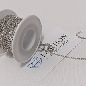May include: A spool of silver rhinestone chain sits next to a piece of the chain and a white card with the text "FASHION - RHINESTONES -" and a diamond logo. The chain is made of small, sparkling rhinestones.