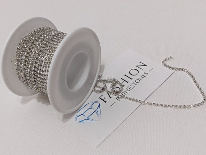 May include: A spool of silver rhinestone chain sits next to a piece of the chain and a white card with the text "FASHION - RHINESTONES -" and a diamond logo. The chain is made of small, sparkling rhinestones.