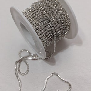 May include: A spool of silver rhinestone chain. The chain is tightly wound around a white plastic spool. A length of the chain extends from the spool, lying loosely on a white surface. The rhinestones sparkle, reflecting light.