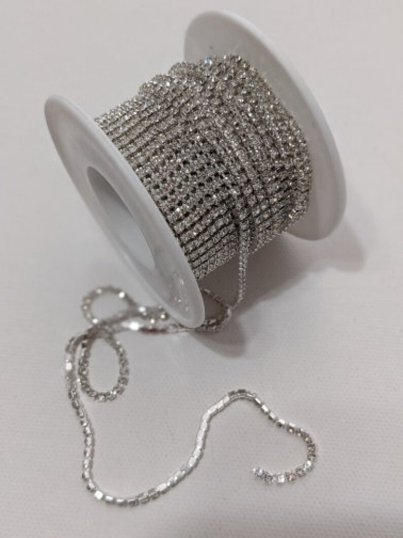 May include: A spool of silver rhinestone chain. The chain is tightly wound around a white plastic spool. A length of the chain extends from the spool, lying loosely on a white surface. The rhinestones sparkle, reflecting light.