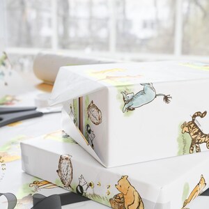 Close-up of a wrapped gift box using Classic Winnie the Pooh-themed wrapping paper, showing detailed illustrations of Pooh, Tigger, and Eeyore. The box rests on a bright white surface with scissors and tape in the background, emphasizing the paper’s smooth glossy texture and charming hand-drawn style.