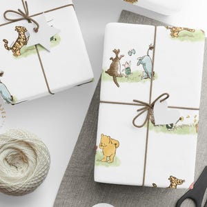 Two wrapped gift boxes tied with brown twine and heart-shaped tags displayed on a light fabric surface. The wrapping paper features soft watercolor illustrations of Winnie the Pooh, Piglet, Eeyore, and Tigger on grassy patches. A spool of white twine and scissors sit nearby for a cozy, handmade aesthetic.