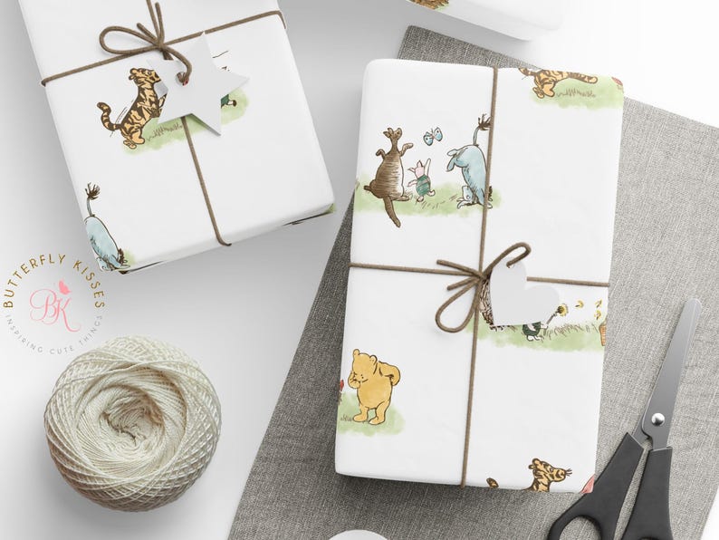 Two wrapped gift boxes tied with brown twine and heart-shaped tags displayed on a light fabric surface. The wrapping paper features soft watercolor illustrations of Winnie the Pooh, Piglet, Eeyore, and Tigger on grassy patches. A spool of white twine and scissors sit nearby for a cozy, handmade aesthetic.