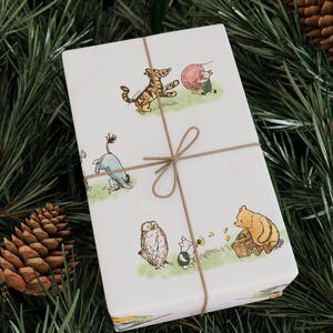 Beautifully wrapped gift in Classic Winnie the Pooh-themed wrapping paper placed among pine branches and pinecones. The hand-drawn illustrations of Pooh, Piglet, Eeyore, and Tigger evoke a nostalgic woodland holiday mood, tied neatly with rustic brown string.