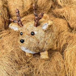 May include: A decorative deer figurine made from natural fibers, featuring brown woven antlers, black eyes, and a black nose. The deer has a light brown body with white accents on the ears and face. The figurine is resting on a bed of similar fibers.