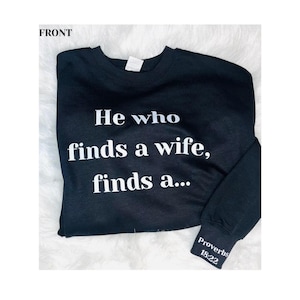 May include: Black sweatshirt with white text: "He who finds a wife, finds a...". The sleeve cuff has the text "Proverbs 18:22". The sweatshirt is displayed on a white, fluffy surface.