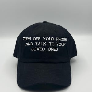 Peut inclure: Casquette de baseball noire avec texte blanc brodé : "TURN OFF YOUR PHONE AND TALK TO YOUR LOVED ONES." La casquette a une visière incurvée et une sangle réglable. Fabriquée en tissu doux et durable.