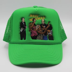 Puede incluir: Gorra de camionero verde brillante con parte trasera de malla y visera curva. La parte delantera presenta un diseño bordado con el texto "JERRY STINGER" sobre un grupo de figuras. Un accesorio vibrante.