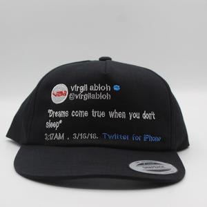 以下が含まれることがあります： 白と青の刺繍が施された黒いスナップバックキャップ。テキストは「virgil abloh @virgilabloh」と「Dreams come true when you don't sleep」で、日付と時刻が記載されています。キャップにはフラットブリムが付いています。