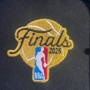 K&ouml;nnte beinhalten: Ein runder gestickter Aufn&auml;her mit einem goldenen Basketball-Motiv, dem schwarzen Schriftzug Finals und der Jahreszahl 2026. Der Aufn&auml;her enth&auml;lt das rot-wei&szlig;-blaue NBA-Logo in der unteren Mitte und ist auf einem schwarzen Stoffuntergrund.
