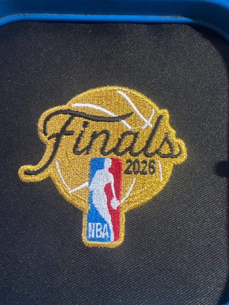 K&ouml;nnte beinhalten: Ein runder gestickter Aufn&auml;her mit einem goldenen Basketball-Motiv, dem schwarzen Schriftzug Finals und der Jahreszahl 2026. Der Aufn&auml;her enth&auml;lt das rot-wei&szlig;-blaue NBA-Logo in der unteren Mitte und ist auf einem schwarzen Stoffuntergrund.