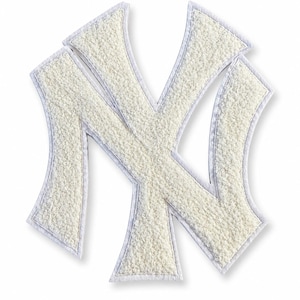 Op de afbeelding: Een witte New York Yankees logo patch met een gestructureerd, pluizig, sherpa-achtig oppervlak. Het iconische NY-embleem is omlijnd met een gladde, strakke witte rand, wat een duidelijk contrast creëert tussen het zachte midden en de randen.