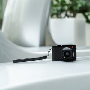 May include: A black digital camera with a black leather strap. The camera has a silver lens with the text "SUMMILUX 1:1.7/28 ASPH" printed on it.