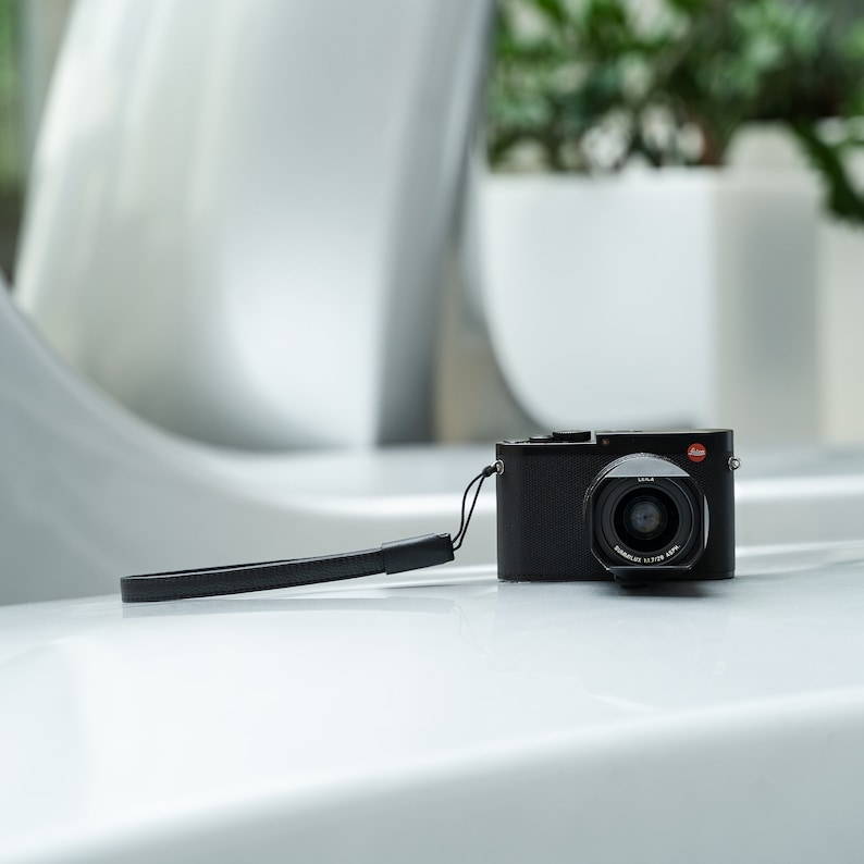 May include: A black digital camera with a black leather strap. The camera has a silver lens with the text "SUMMILUX 1:1.7/28 ASPH" printed on it.
