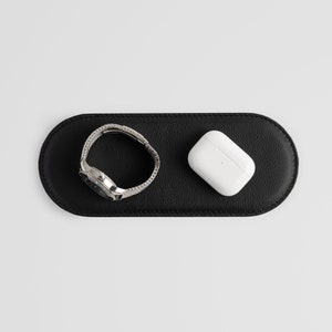Può includere: Un tappetino di ricarica in pelle nera con un orologio argentato e una custodia di ricarica wireless bianca.