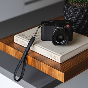 May include: A black camera with a silver lens sits on top of a book titled "The Monocle Guide to Good Business". A black leather camera strap is attached to the camera.