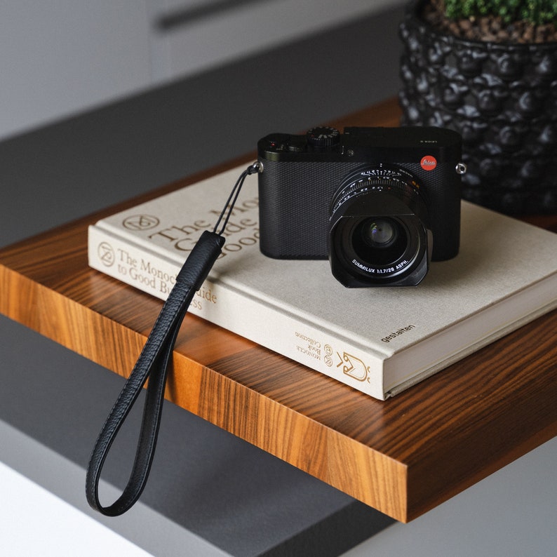 May include: A black camera with a silver lens sits on top of a book titled "The Monocle Guide to Good Business". A black leather camera strap is attached to the camera.