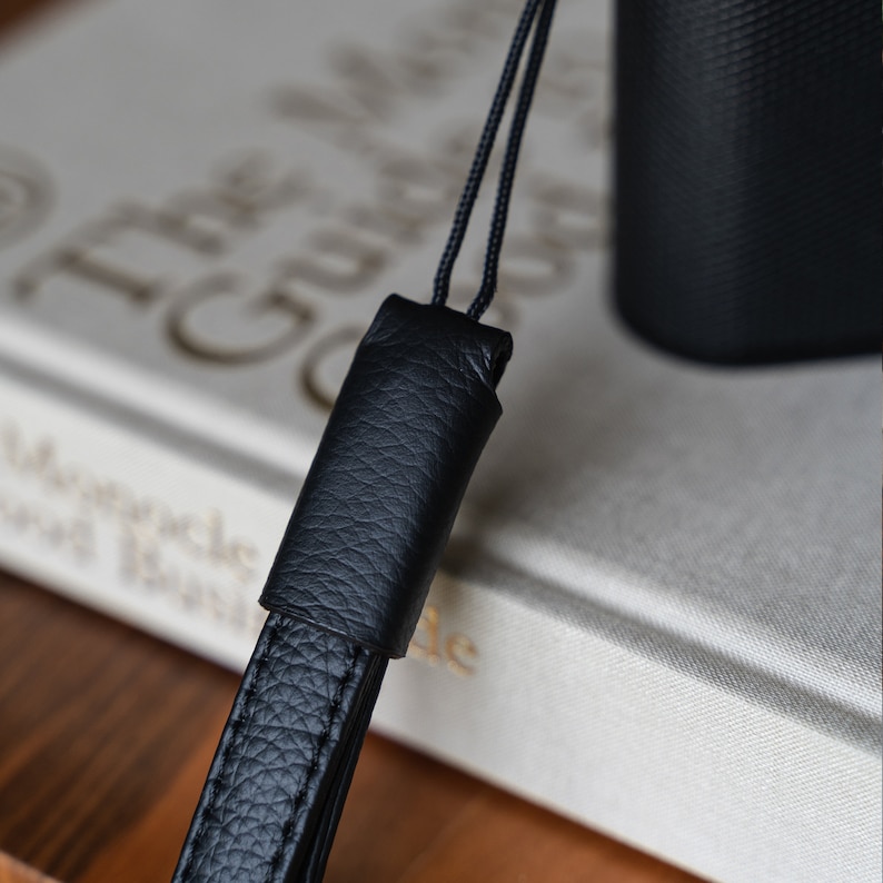 May include: Close-up of a black leather camera strap with a textured surface. The strap has a loop at the end for attaching to a camera.