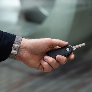 May include: A hand holding a black leather key holder with a silver key attached. The key holder has a silver button on the side. The hand is wearing a silver metal bracelet.