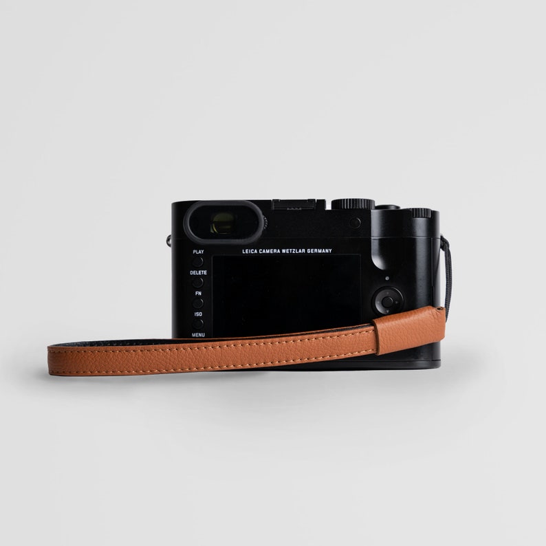 May include: A black Leica camera with a brown leather strap. The camera has a screen and buttons on the back, and a lens on the front. The text "LEICA CAMERA WETZLAR GERMANY" is printed on the camera.