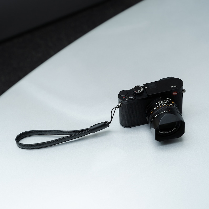 May include: A black camera with a black leather strap. The camera has a silver lens and a silver dial on the top.