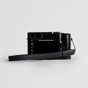May include: A black Leica camera with a black leather strap. The camera has a screen on the back and buttons for play, delete, fn, and iso.