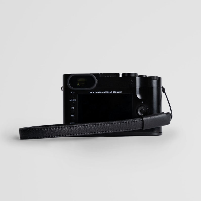 May include: A black Leica camera with a black leather strap. The camera has a screen on the back and buttons for play, delete, fn, and iso.