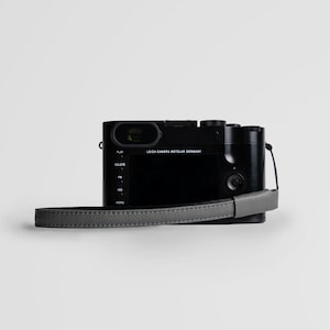 May include: A black Leica camera with a gray leather strap. The camera has a screen on the back and buttons for play, delete, fn, iso, and menu.