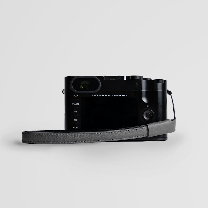 May include: A black Leica camera with a gray leather strap. The camera has a screen on the back and buttons for play, delete, fn, iso, and menu.