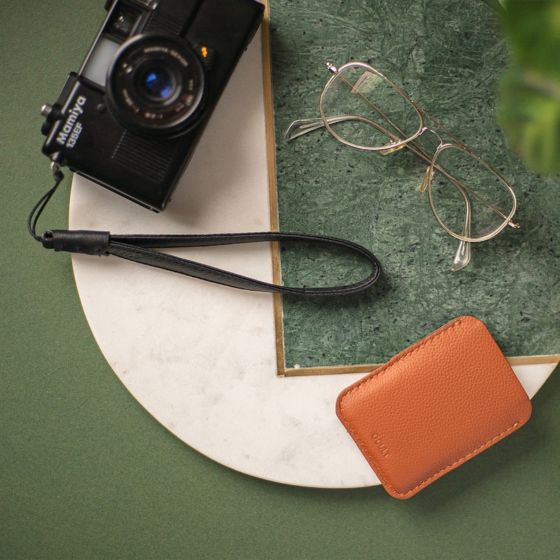 May include: A vintage camera with a black leather strap, a pair of gold-rimmed glasses, and a brown leather wallet on a white marble surface with a green background.