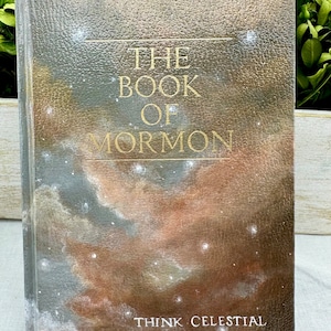 Puede incluir: El Libro de Mormón con una portada con temática de galaxia. La portada es de cuero marrón texturizado con un diseño de galaxia pintado en tonos grises, marrones y blancos. El título "El Libro de Mormón" está escrito en letras doradas. La parte inferior de la portada dice "Think Celestial" en letras blancas.