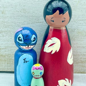May include: A set of three wooden peg dolls, one blue with a Stitch character design, one green with a pink bow and a stitched design, and one red with a white floral design and black hair.