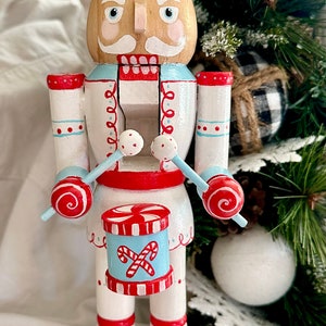 Puede incluir: Un cascanueces de madera vestido con un atuendo de temática de bastón de caramelo blanco y azul, sosteniendo baquetas y un tambor de bastón de caramelo. El cascanueces lleva un sombrero a rayas azul y blanco con una corona roja.