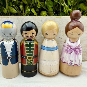 May include: Four hand-painted wooden peg dolls. The dolls are painted to resemble characters, including a mouse, a nutcracker, a woman in a cream dress with a blue sash, and a woman in a lavender dress.