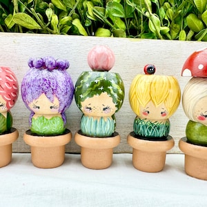 Puede incluir: Seis muñecas de madera pintadas a mano con caras y cabello, cada una con un sombrero de maceta diferente. Las muñecas están dispuestas en fila sobre una superficie blanca.