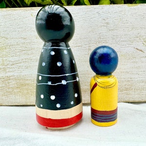 K&ouml;nnte beinhalten: Zwei Holz-Steckpuppen, eine mit einem schwarz-wei&szlig;en Polka-Dot-Kleid und einem rot-wei&szlig;en Streifen am unteren Rand, und die andere mit einem gelben Hemd und einem blau-roten Streifen am unteren Rand.