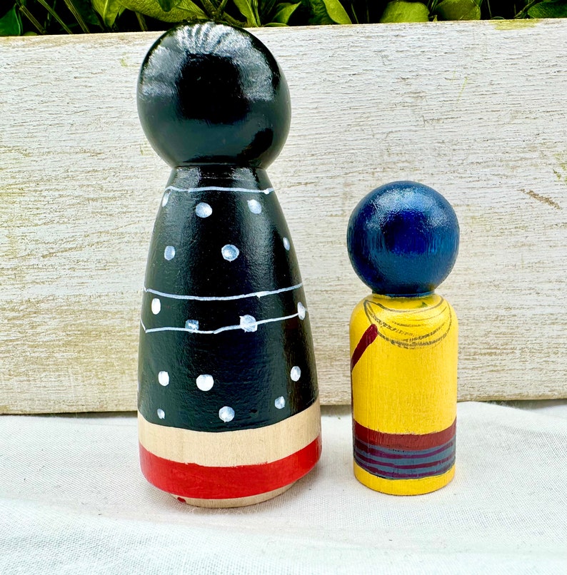 K&ouml;nnte beinhalten: Zwei Holz-Steckpuppen, eine mit einem schwarz-wei&szlig;en Polka-Dot-Kleid und einem rot-wei&szlig;en Streifen am unteren Rand, und die andere mit einem gelben Hemd und einem blau-roten Streifen am unteren Rand.