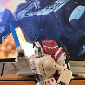 May include: A white and red robot toy with a blue sword, standing in front of a computer screen displaying a blue and gray robot. The robot toy has a red and white jet pack and a white helmet with a blue visor.