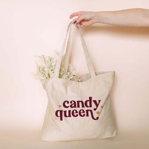 May include: A beige canvas tote bag with the words "candy queen" in burgundy script and star accents. The bag is filled with white flowers and held by a person.