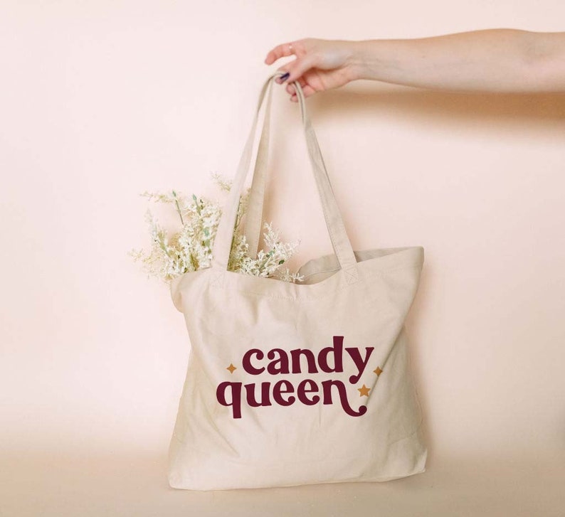 May include: A beige canvas tote bag with the words "candy queen" in burgundy script and star accents. The bag is filled with white flowers and held by a person.