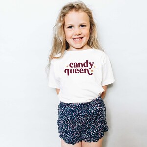May include: A young girl wearing a white t-shirt with the words "candy queen" in burgundy script and gold stars. She is also wearing blue and white patterned shorts. The girl is smiling.