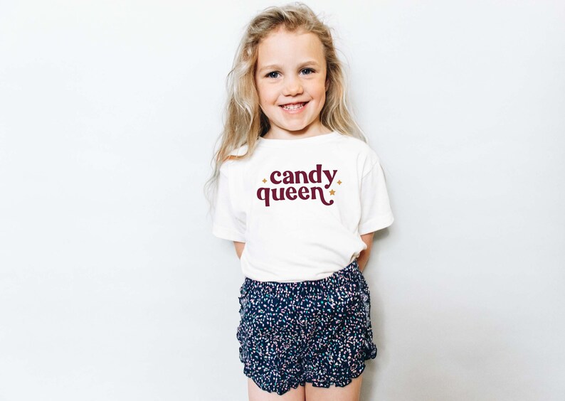 May include: A young girl wearing a white t-shirt with the words "candy queen" in burgundy script and gold stars. She is also wearing blue and white patterned shorts. The girl is smiling.